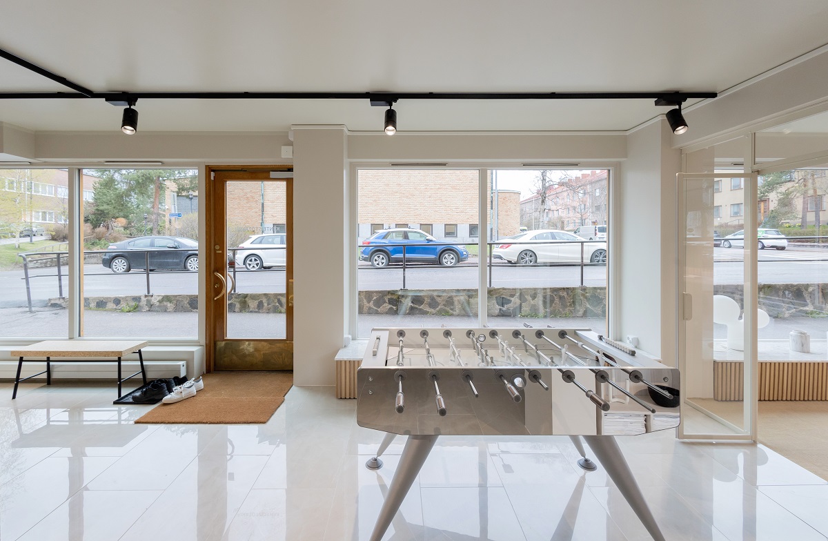 Nykyaikainen sisätila, jossa on suuret ikkunat esittelemässä ulkona pysäköityjä autoja, jossa on jalkapallopöydän kiiltävällä lattialla. Penkki, jossa on kenkäline ja ovimatto, sijaitsee noin LED-kiskovalaistuksen alla, korostaen huoneen modernia muotoilua. Tämä kaikki löytyy LedStore.fi:n laajasta valikoimasta LED-valaistusratkaisuja kotisi sisustukseen.