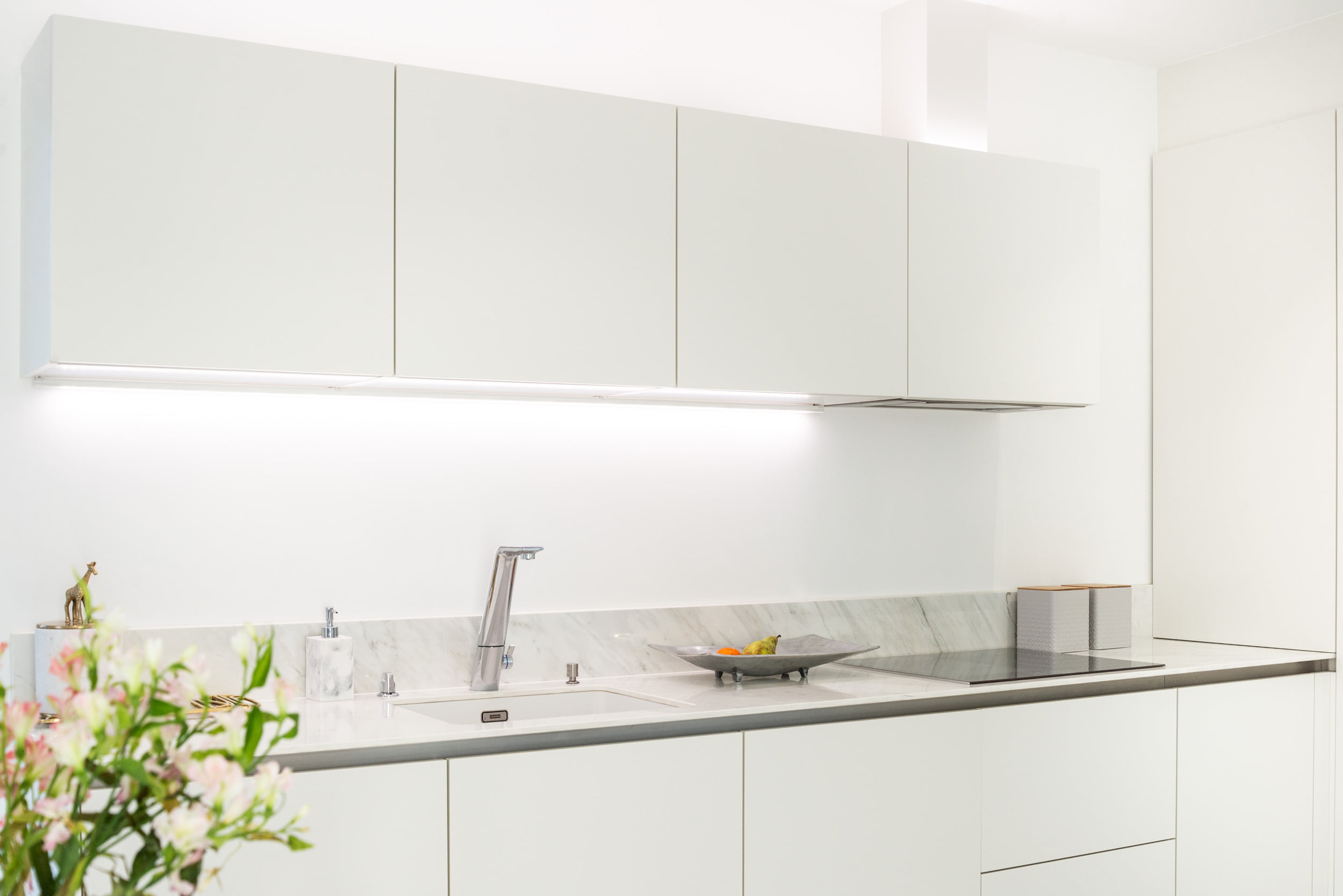 Tyylikäs keittiö, jossa on puhtaat valkoiset kaapistot, moderni marmoritaso ja ruostumattomasta teräksestä valmistettu hana. Työtason alle asennetut LED-valaisimet korostavat työtilaa tulemista. Dekoratiivinen kasvi ja hedelmäkulho tuovat lisää ilmettä tasolle. Voit löytää sopivat LED-valaistusratkaisut LedStore.fi-verkkokaupasta, josta saat laadukkaita LED-tuotteita keittiösi ilmeen täydentämiseksi.