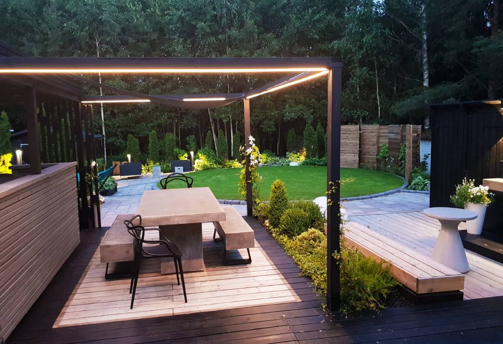 Moderni ulkoterassi pergolalla, jossa on sisäänrakennettu LED-valaistus, betonipöytä, penkkejä puisella kannella; rehevä puutarha ja hoidettu nurmialue.