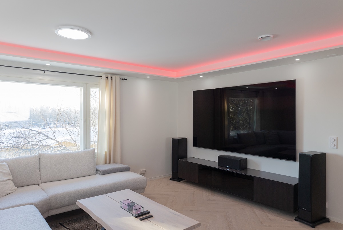 Moderni olohuone täynnä tyylikkäätä tunnelmaa. Suuri sohva kutsuu rentoutumaan, ja vaalea puinen sohvapöytä tuo tilaan lämpöä. Seinällen taulutelevisio, jossa on LED-taustavalaistus, lisää huoneeseen modernia ilmettä. Vahvista elämyksiä lattiakaiuttimilla ja nauti punaisesta LED-korostusvalaistuksesta katon rajassa. Iso ikkuna päästää sisään päästä luonnonvaloa, tehden tilasta avaran ja valoisan. Kaikki nämä elementit yhdessä luovat täydellisen ympäristön LedStore.fi:n monipuolisilla LED-valaistusratkaisuilla.