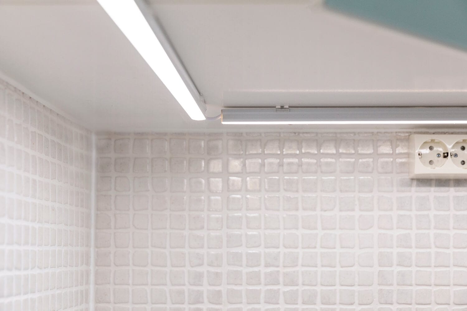 Moderni keittiön backsplash valkoisilla teksturoiduilla laatoilla, jotka valaisevat kaapin alla olevalla lineaarisella LED-valolla; lempeä hehku korostaa seinässä olevaa pistorasiaa.