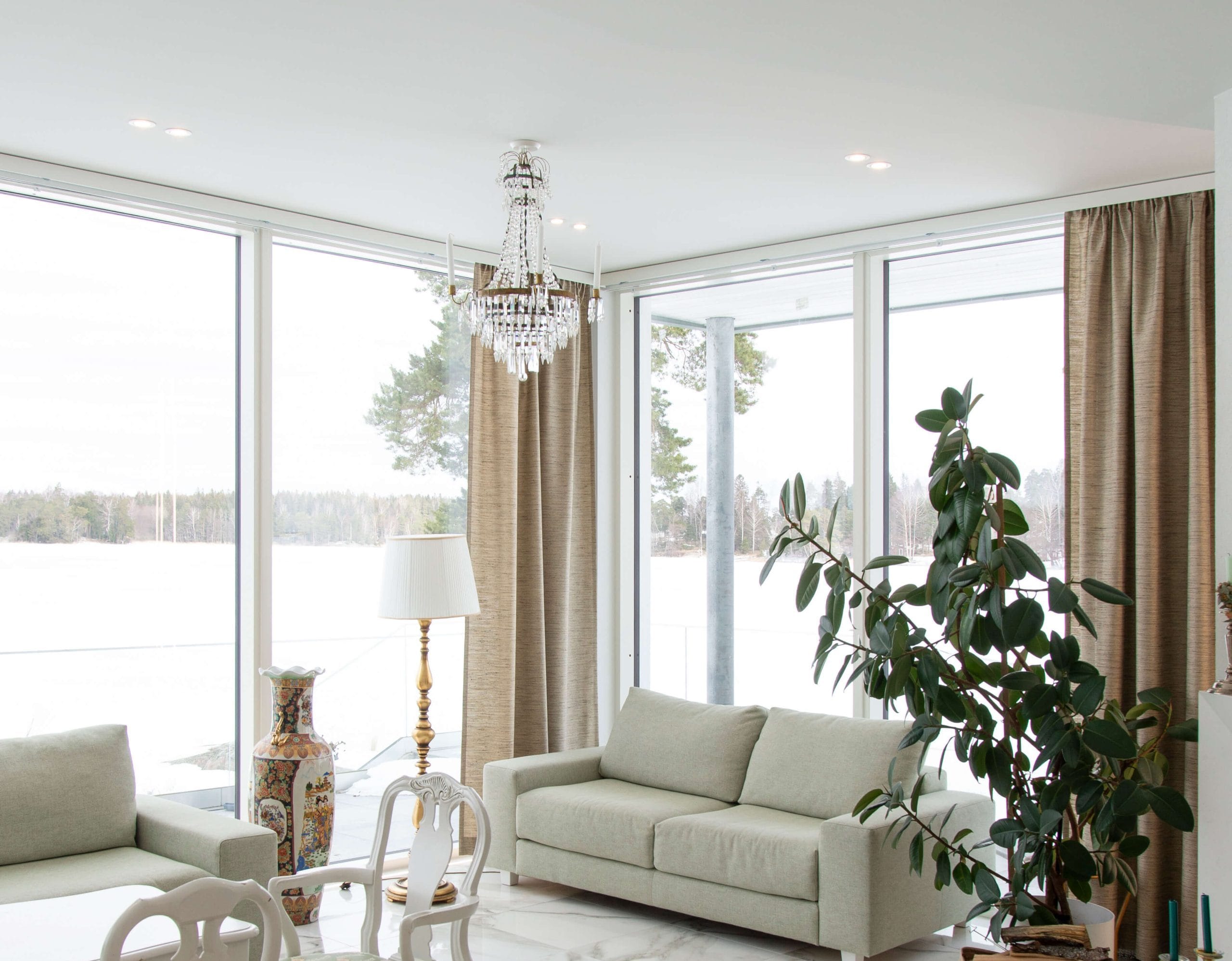 Valoisassa olohuoneessa, jossa on suuret ikkunat lumiseen maisemaan, on valkoinen sohva, suuri viherkasvi ja tyylikäs kattokruunu, jonka LED-valaisimet tuovat esiin sen kauneuden. Beiget verhot täydentävät koristeellista maljakkoa ja lattiavalaisinta, kun taas ruokapöytä valkoisine tuoleineen pilkahtelee taustalla. Tutustu LedStore.fi:n laajaan LED-valaisinvalikoimaan ja luo kotiisi yhtä upea valaistusratkaisu!