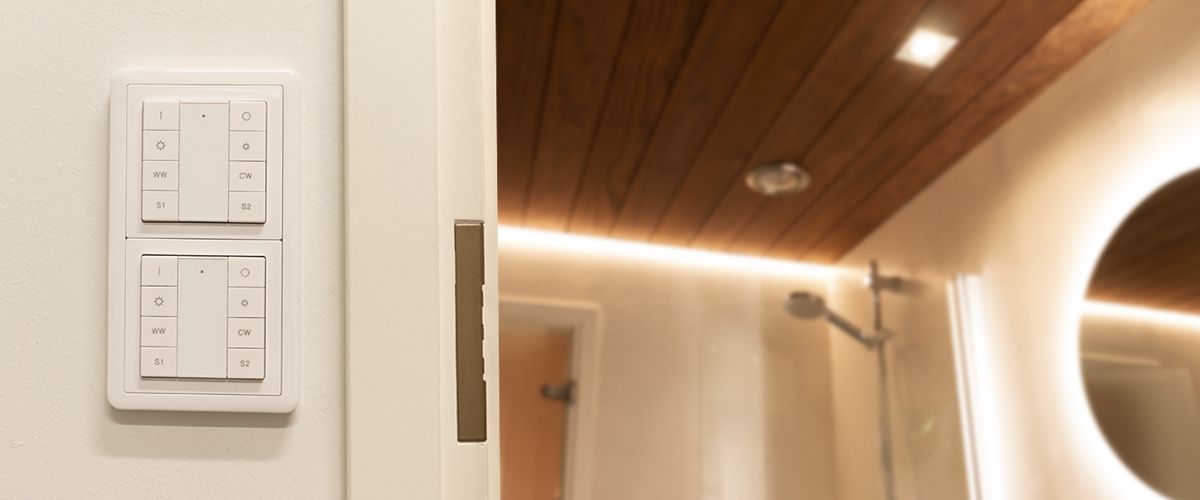 Kylpyhuone, jossa on puiset kattopaneelit ja pehmeä LED-valaistus. Tilasta löytyy myös kätevä walk-in-suihku sadesuihkupäällä. Läheisellä seinällä on kaksi modernia valokytkimen paneelia, ohjataan näppäimillä. Tyylikkään kokonaisuuden kruunaa pyöreä led-valaistu peili.